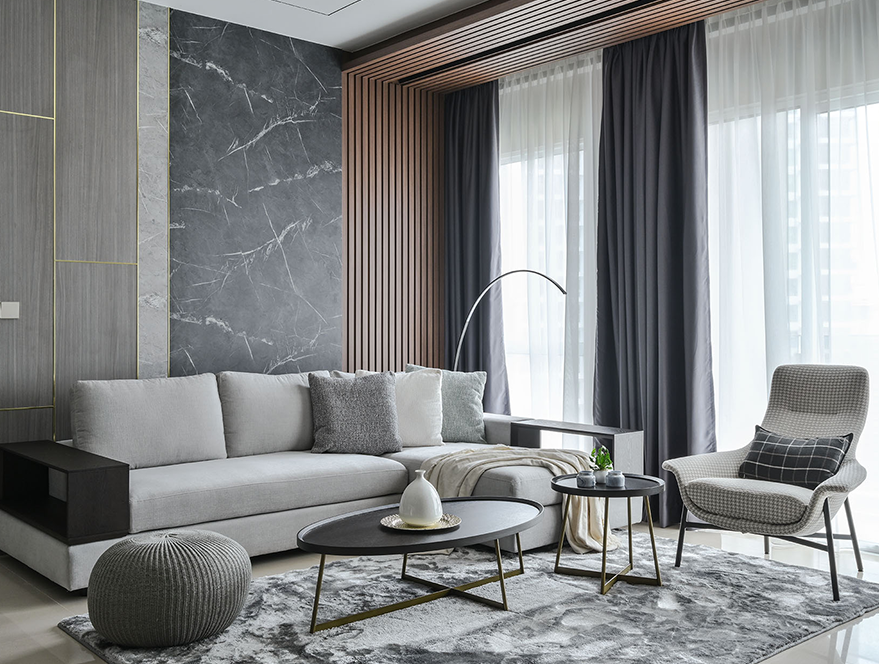 A modern living room with a light grey sectional sofa, black coffee table, patterned armchair, marble-effect wall paneling, and grey window curtains.