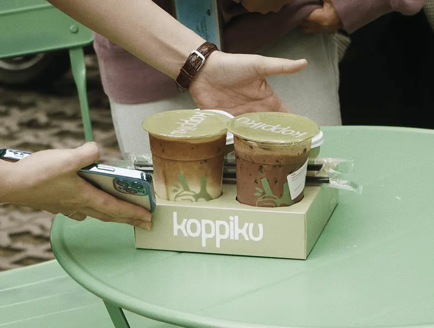 Two iced Koppiku drinks in a cardboard carrier resting on a light green outdoor table, with a person holding a smartphone nearby.