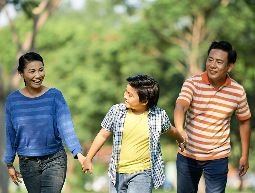 A happy Asian family of three, including a mother, father, and son, walking through a park while holding hands.