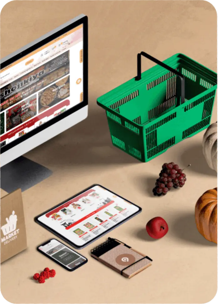 A still life arrangement illustrating online grocery shopping. A desktop monitor displays a website labeled "shokikiya," a tablet and smartphone show a shopping app, and a large green plastic shopping basket sits beside the monitor. Produce like grapes, an apple, and pumpkins are scattered nearby.